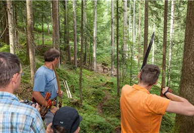 Eine Gruppe von Personen steht im Wald und hält Bögen in der Hand. Sie beobachten aufmerksam die Umgebung zwischen den Bäumen.