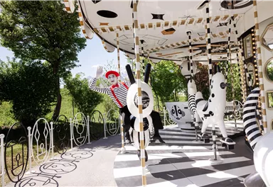 A carousel with a cheerful mime in a striped outfit. The surroundings are green and sunny, featuring a creative design of the attraction.