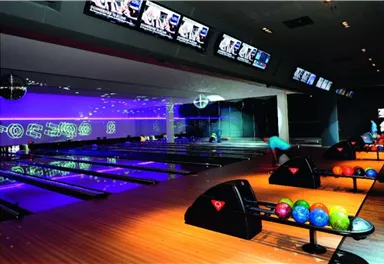 A modern bowling alley with colorful bowling balls and well-lit lanes. TVs are mounted on the wall, showing sports broadcasts.