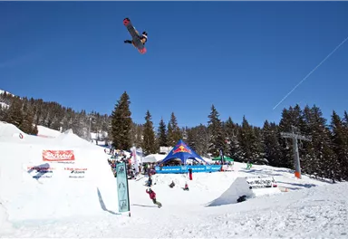Ein Snowboarder springt hoch in die Luft über ein Snowpark-Set-up im Schnee. Im Hintergrund sind Bäume und ein blauer Himmel zu sehen.