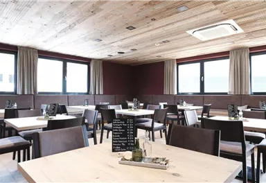 A modern restaurant with light wooden furniture and large windows. The tables are neatly set and the space feels inviting and spacious.