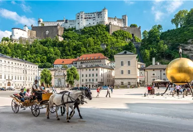 Ein schöner Platz in Salzburg mit einer Pferdekutsche und historischer Architektur. Im Hintergrund sieht man eine beeindruckende Festung auf einem Hügel.