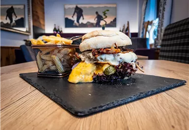 A juicy burger with cheese and fresh vegetables is served on a black platter. Accompanied by crispy fries in a small basket.