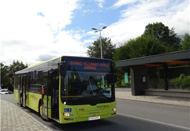 A yellow bus stops at a bus stop. In the background, there are trees and a partly cloudy sky.