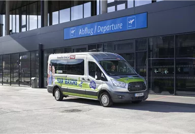 A city bus is standing in front of a modern airport building. Above the bus, the sign "Abflug / Departure" is visible.