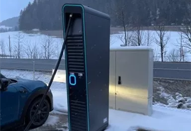 Eine Ladestation für Elektroautos steht auf einem schneebedeckten Parkplatz. Im Hintergrund sind Bäume und eine winterliche Landschaft zu sehen.