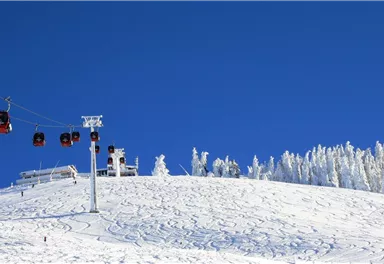 Eine verschneite Berglandschaft mit einer Seilbahn und blauem Himmel. Die Bäume sind von Schnee bedeckt und die Umgebung strahlt Winteridylle aus.