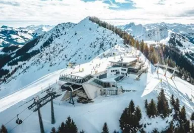 Eine schneebedeckte Berglandschaft mit Skiliften und einer modernen Bergstation. Im Hintergrund sind majestätische Berggipfel und ein klarer Himmel zu sehen.