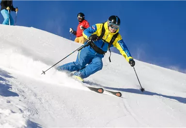A skier in colorful clothing is skiing down the snow-covered slope. In the background, more skiers and a clear blue sky can be seen.