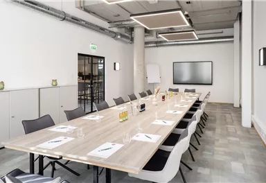 Ein moderner Besprechungsraum mit einem langen Tisch und Stühlen. An der Wand hängt ein großer Bildschirm, und es gibt verschiedene Getränke und Schreibmaterialien auf dem Tisch.
