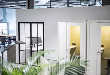 Ein modernes Büro mit gläsernen Besprechungsräumen und geschlossenen Telefonkabinen. Im Vordergrund sind grüne Pflanzen zu sehen, die eine angenehme Atmosphäre schaffen.