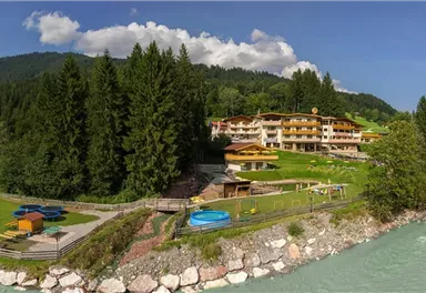 A picturesque landscape with a hotel by the river and green meadows. In the background, majestic mountains rise under a blue sky.