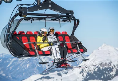 A cable car with two skiers sitting in the mountains. In the background, snow-covered peaks and a clear blue sky can be seen.