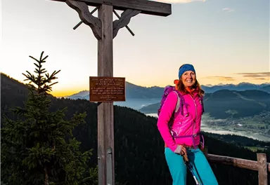 A hiker stands at a wooden cross on a mountain. In the background, the mountains and a clear sky can be seen.