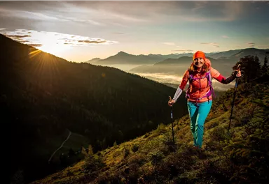 A woman hikes in the mountains at sunrise. The landscape is green and picturesque with a clear sky in the background.