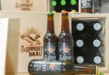 Ein Holzstapel mit Bierflaschen der Marke "Sunnseit Bräu" und einem Logo. Umgeben von einigen Hopfen und Holzboxen.