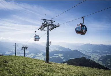 A cable car travels over a green meadow with mountains in the background. The sky is clear and blue.