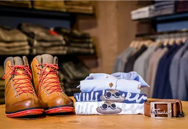 A pair of brown hiking boots with red laces is sitting on a wooden table. Next to it are clothes and a belt in attractive colors.