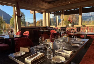 Ein elegantes Restaurant mit Holzdekor und roten Stühlen. Der Tisch ist festlich gedeckt mit Gläsern und Geschirr, und große Fenster bieten einen Blick auf die Landschaft.