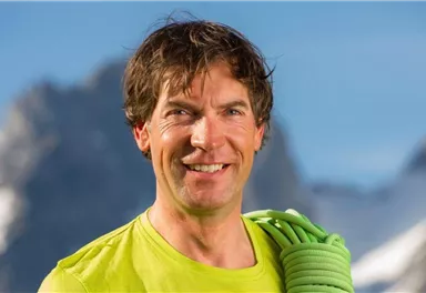 A sporty man with short hair smiles at the camera. He is wearing a green shirt and is holding a climbing rope in his hand, while the mountains are visible in the background.