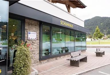 Ein moderner Laden mit dem Namen "SCHUHHAUS" in einer naturnahen Umgebung. Vor dem Geschäft stehen Bänke und es gibt geringen Platz für Grünanlagen.