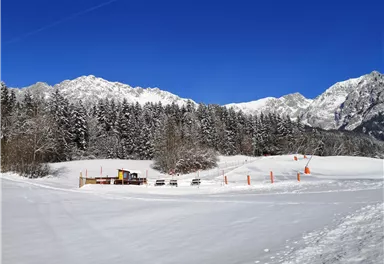Eine verschneite Landschaft mit hohen Bergen im Hintergrund. Einige Schaufel- und Sitzgelegenheiten sind sichtbar, umgeben von schneebedeckten Bäumen.