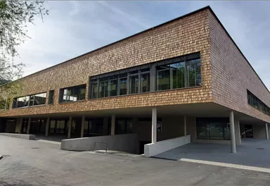 A modern building with wooden cladding and large windows. It is located in a rural area with a green backdrop.