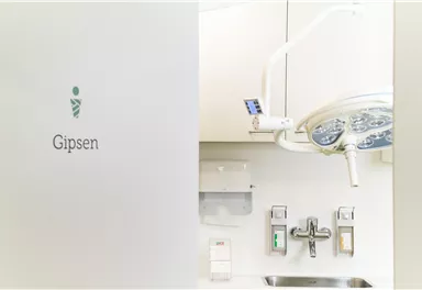 Ein moderner Behandlungsraum mit einem zahnärztlichen Stuhl und einer Deckenleuchte. An der Wand ist ein Schild mit der Aufschrift "Gipsen" zu sehen.