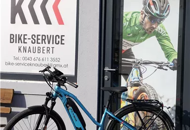 Ein blaues E-Bike steht vor einem Geschäft mit dem Namen "Bike-Service Knaubert". Im Hintergrund ist ein großes Bild eines Radfahrers zu sehen.