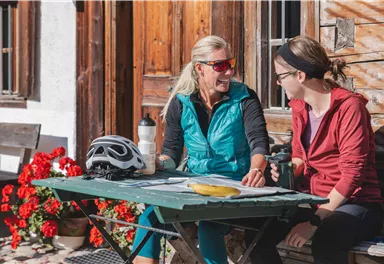 Zwei Frauen sitzen an einem Tisch im Freien und unterhalten sich. Auf dem Tisch liegt ein Fahrradhelm, eine Wasserflasche und eine Banane.