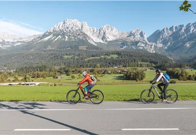 Zwei Radfahrer fahren entlang einer Straße auf einer weiten grünen Wiese. Im Hintergrund sind beeindruckende Berge und ein klarer blauer Himmel zu sehen.