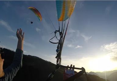 Ein Paraglider schwebt über einer malerischen Landschaft bei Sonnenuntergang. Eine Person streckt die Hand aus und beobachtet den Paraglider.