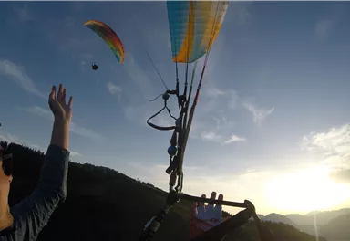 Ein Paraglider schwebt über einer malerischen Landschaft bei Sonnenuntergang. Eine Person streckt die Hand aus und beobachtet den Paraglider.