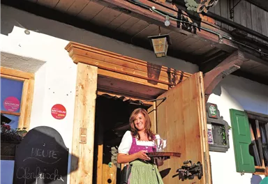 Eine Frau in traditioneller Tracht steht vor einem Gasthaus und hält ein Tablett mit Getränken. Die Fassade des Hauses ist mit Blumen geschmückt.
