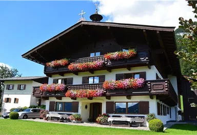 Ein traditionelles alpenländisches Haus mit farbenfrohen Blumenkästen an den Balkonen. Umgeben von einer gepflegten Rasenfläche und Bäumen.