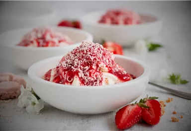 A bowl of ice cream, topped with strawberry sauce and coconut flakes. Fresh strawberries are placed beside it.