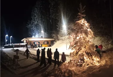 A festively lit fir tree stands in the snow as people gather in a winter mood. In the background, a cozy cabin with lights can be seen.