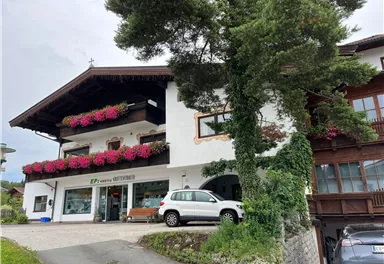 Ein traditionelles Gebäude mit Balkon und blühenden Pflanzen. Vor dem Haus steht ein weißes Auto und es ist von Bäumen umgeben.