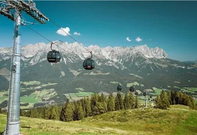 Eine Seilbahn fährt über eine grüne Landschaft mit Bergen im Hintergrund. Der Himmel ist klar und blau.