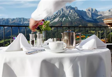 A set dining table with a teapot and a flower vase in the mountains. The surroundings are picturesque with a clear view of the mountains.