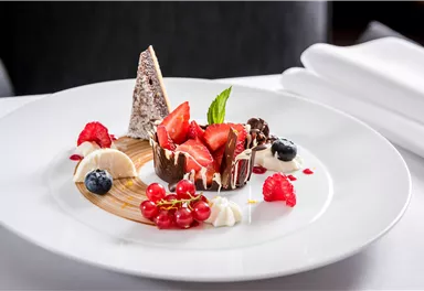 An artistically arranged dessert on a white plate. It consists of fresh fruits, chocolate, and a decorative cream.