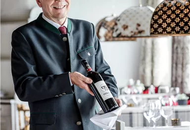 A smiling waiter in classic attire holds a bottle of wine. In the background, an elegantly set table can be seen in an appealing restaurant.