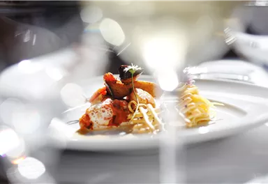 An elegant dish on a white plate, decorated with fresh ingredients. In the background, blurred glasses are visible, creating an upscale atmosphere.