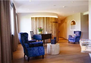 An elegant reception area with blue armchairs and a round wooden table. In the background, there is a wood paneling with plants.