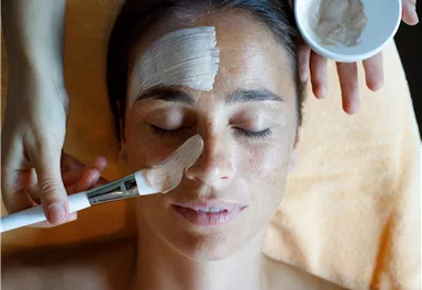 A woman is relaxed on a lounge chair while her face is being treated with a facial mask. One hand holds a brush to apply the mask, while the other hand holds a bowl with productive contents.