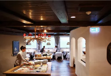 A cozy restaurant with a rustic wooden ceiling and an elegant atmosphere. Some guests are enjoying their meals while the staff prepares the table.