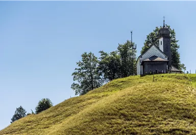 A small church stands on a gentle hill, surrounded by green trees. The sky is clear and blue.