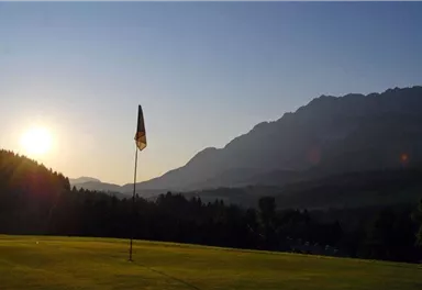 Ein ruhiger Golfplatz mit einer Fahne, umgeben von Bergen. Die Sonne geht hinter den Hügeln auf und schafft eine friedliche Atmosphäre.