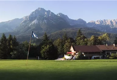 Ein schöner Golfplatz mit einem Flaggenstock im Vordergrund. Im Hintergrund sind majestätische Berge und ein typisches alpenländisches Haus zu sehen.