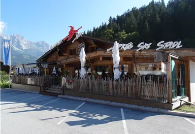 A traditional restaurant in a picturesque setting with wooden paneling and sun umbrellas. In the background, trees and mountains are visible.